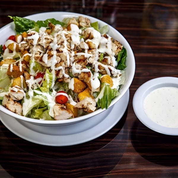 A large chicken Caesar salad with croutons, cherry tomatoes, and dressing, with a side of ranch.